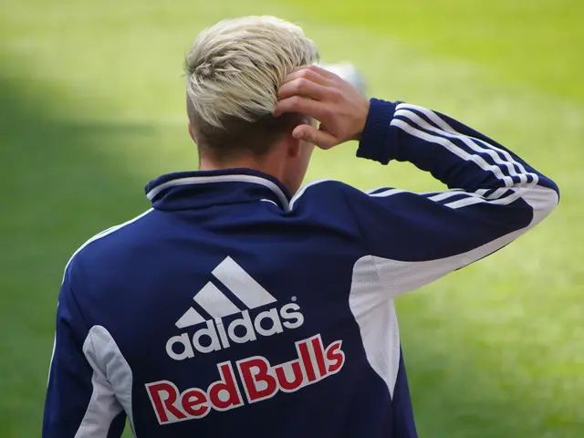 The image shows a man in a blue and white jacket standing on top of a soccer field, with a blurred...