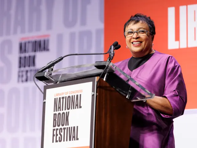 The image shows a woman standing at a podium in front of a screen, wearing a purple dress and...