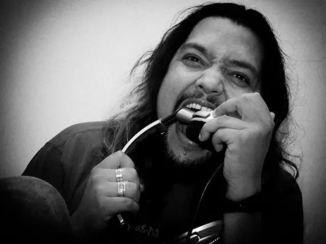 The image shows a man with long hair and a beard singing into a microphone in front of a wall. He...