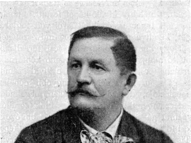 The image shows a black and white photo of a man with a mustache wearing a blazer and a bow tie. At...