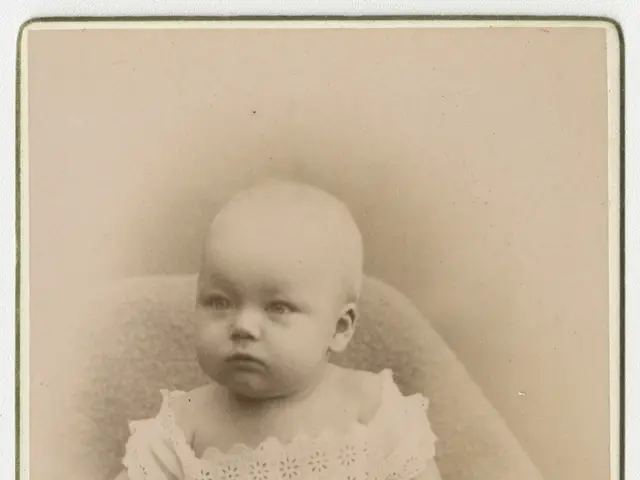 The image shows an old photo of a baby sitting on a blanket, with text at the bottom. The baby has...