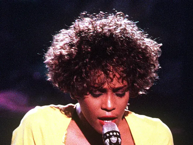 The image shows Whitney Houston performing at the Grammys, wearing a yellow dress and holding a...