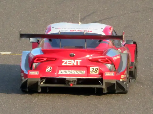 The image shows a red sports car driving on a race track. The car is adorned with text and numbers,...