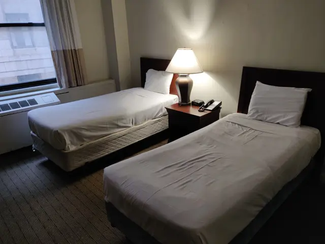 The image shows a hotel room with two beds, each with white bed sheets and pillows, a nightstand...