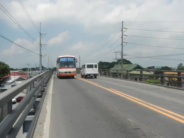 The image shows two buses driving across a bridge with railings on either side. In the background,...