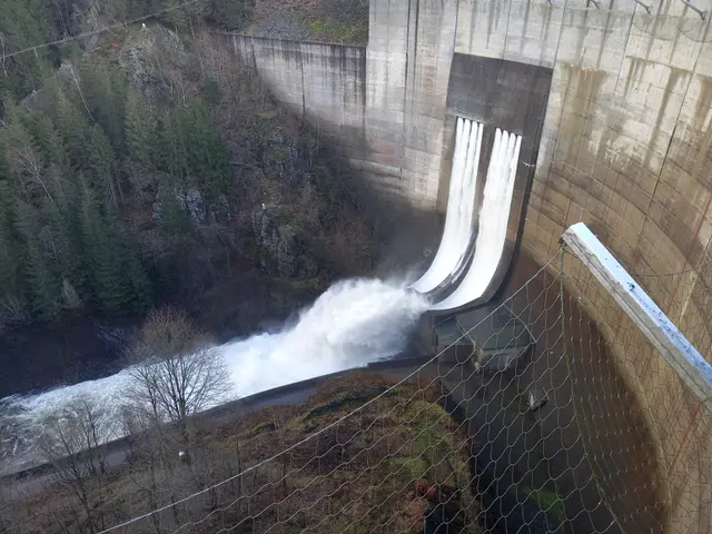 The image shows a large dam with water coming out of it, surrounded by a fence and trees on the...