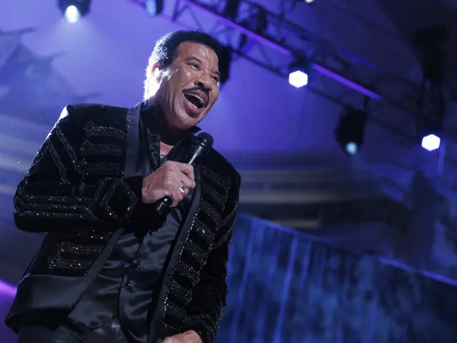 The image shows Lionel Richie performing at the Grammys, wearing a black dress and holding a...