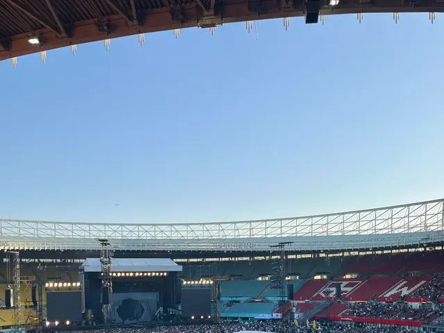 The image shows a large crowd of people sitting in a stadium, with some standing, surrounded by...