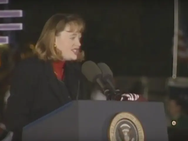 The image shows a woman standing at a podium in front of a crowd, with a logo on the podium and...