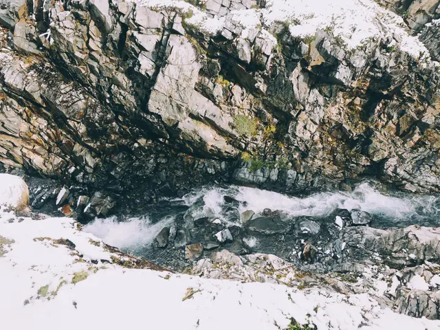The image shows a small stream running through a rocky mountain side covered in snow. The water is...