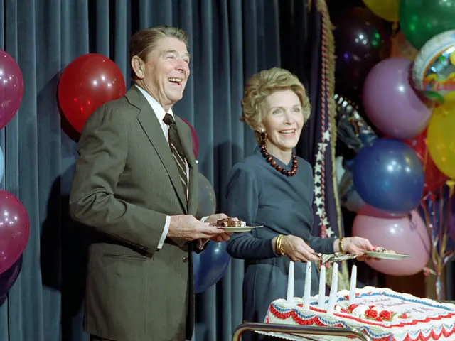 The image shows a man and woman standing next to each other in front of a table with a cake adorned...