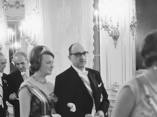 The image shows a man and woman in formal attire walking down a hallway, surrounded by a group of...