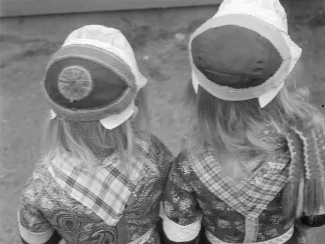The image shows two young girls in traditional Bavarian dresses and hats standing next to each...