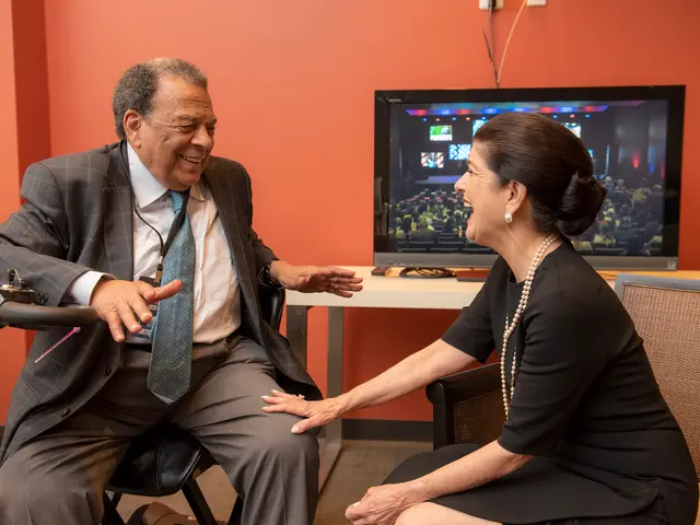 The image shows a man and woman sitting in chairs talking to each other in front of a television on...