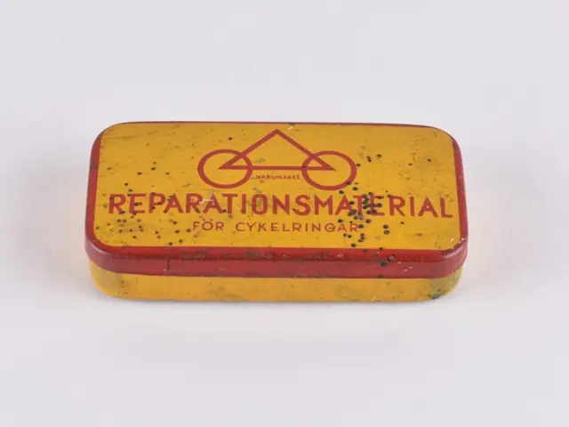 The image shows a small yellow and red tin box with the words "Reparationsmaterial" written on it,...