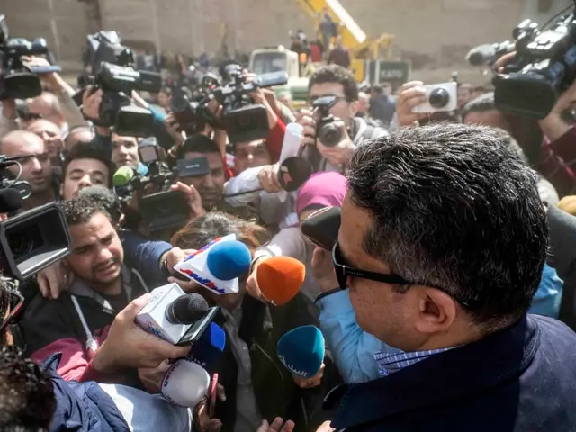 The image shows a man in a blue jacket surrounded by reporters, some of whom are holding cameras...