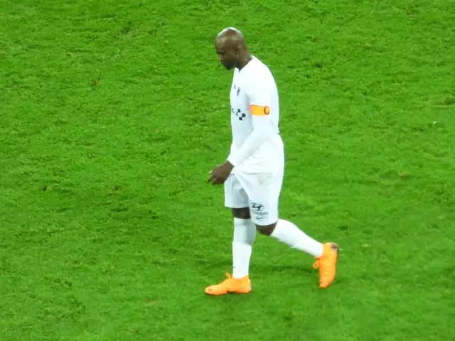 The image shows a soccer player walking across a lush green field, wearing a white t-shirt, white...
