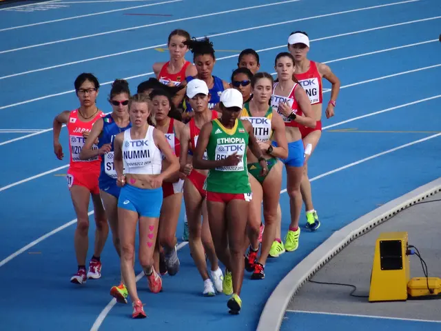 The image shows a group of women running on top of a track, with a person standing on the right...