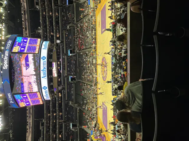 The image shows a basketball game being played in a large arena at night. There are people sitting...