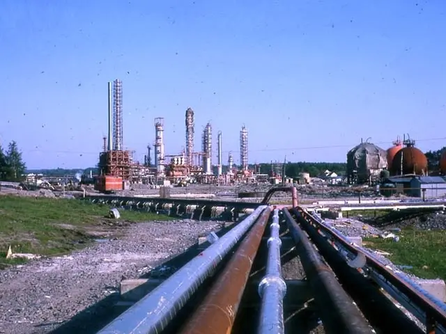 The image shows an oil refinery with a large number of pipes in the foreground, surrounded by...