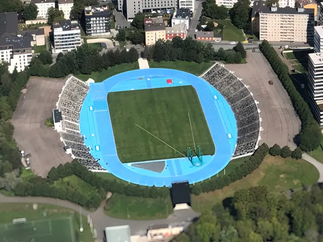 The image shows an aerial view of a large stadium in the middle of a city, surrounded by buildings,...