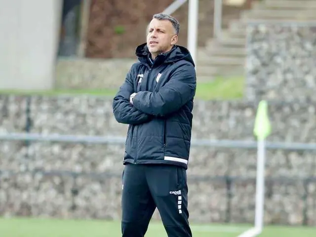 The image shows a man standing on top of a soccer field with his arms crossed, wearing a jacket and...