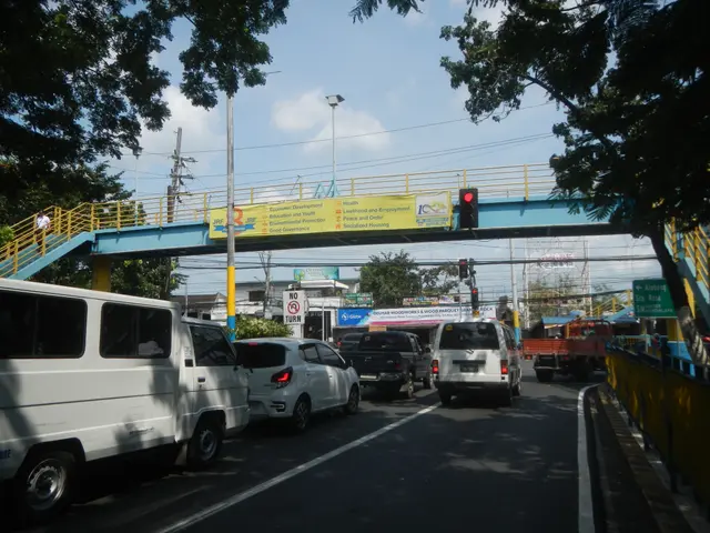 The image shows a busy street with cars and trucks driving down it, a bridge with railings and...