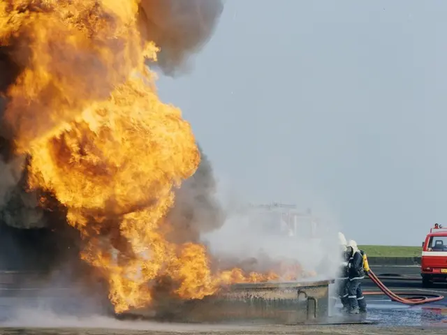 The image shows a fire truck engulfed in flames on the side of a road, with two people wearing...