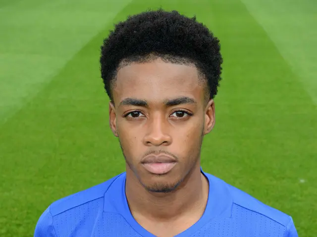 The image shows a young man in a blue t-shirt standing on top of a soccer field. He is wearing a...
