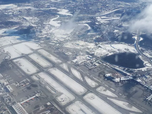 The image shows an aerial view of an airport with snow on the ground, buildings, roads, bridges,...