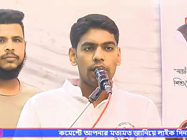 The image shows two men standing side by side in front of a TV screen. The man on the right is...