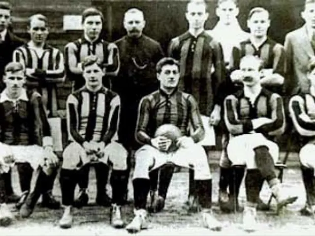 The image shows a black and white photo of a soccer team, with some standing and some sitting, in...