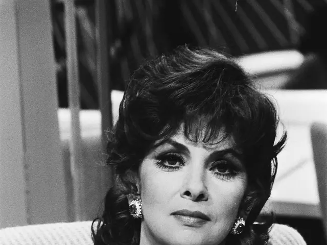 The image shows Elizabeth Taylor sitting on a chair with a microphone in front of her. She is...