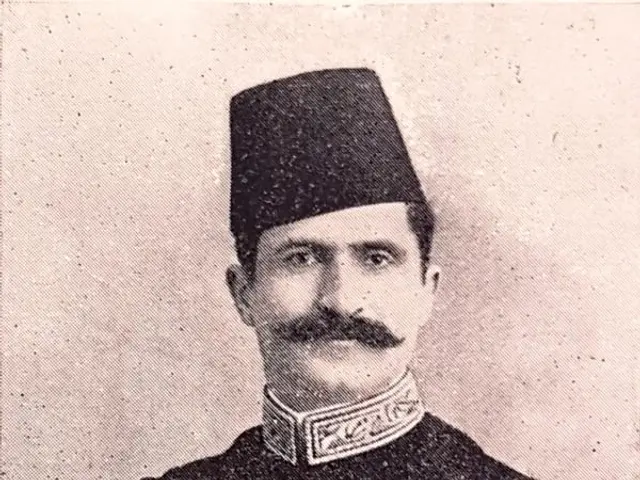 The image shows an old black and white photo of a man in uniform, wearing a cap and adorned with...