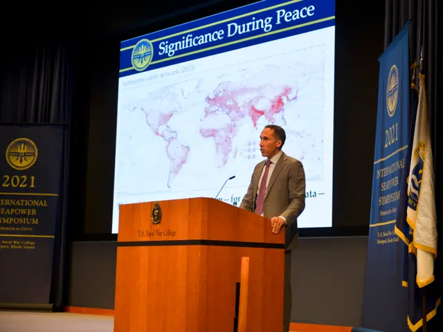 The image shows a man standing at a podium in front of a projection screen, wearing a blazer, tie,...