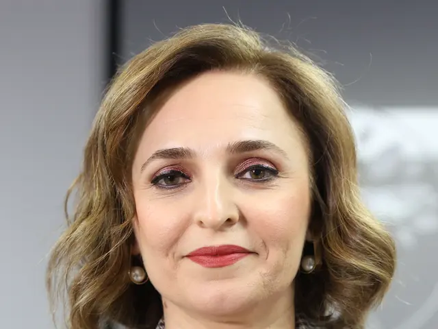 The image shows a woman in a black top and blazer speaking into microphones in front of a blurred...