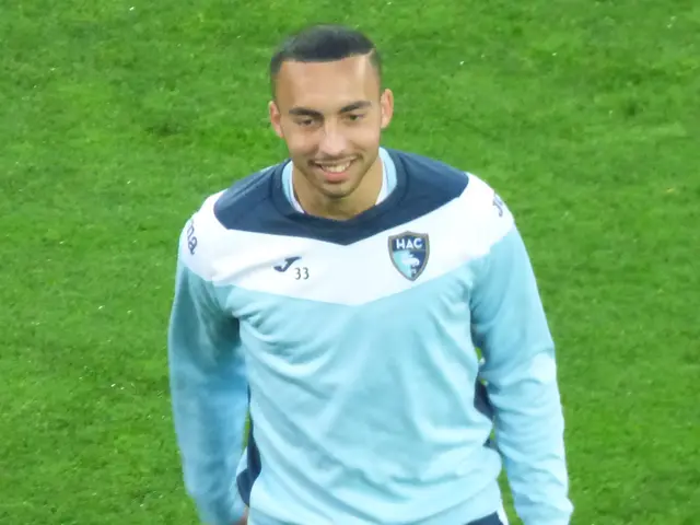 The image shows a man in a blue shirt and black pants walking on a soccer field, with a smile on...