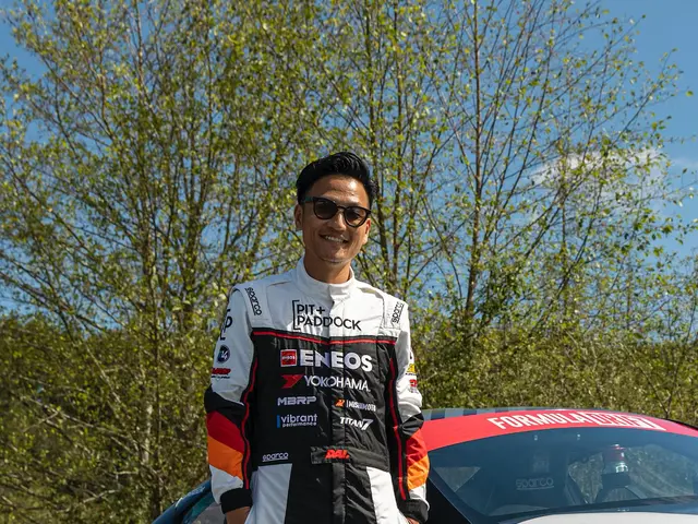 The image shows a man standing next to a race car on a road surrounded by trees and a clear blue...