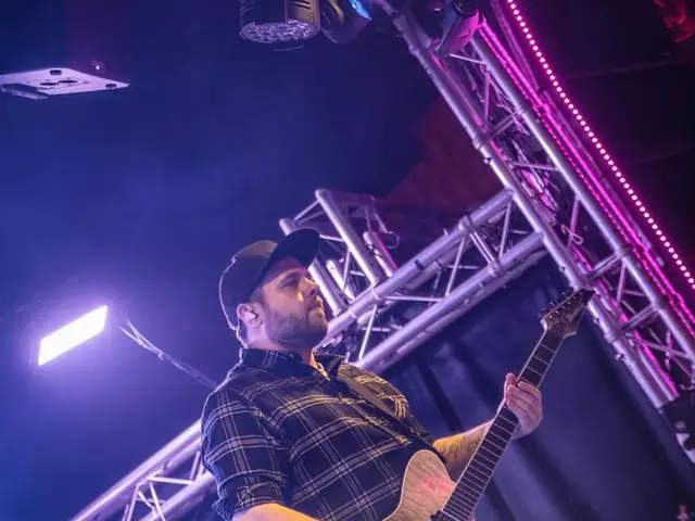The image shows a man playing a guitar on stage at a concert, wearing a cap and surrounded by mics...