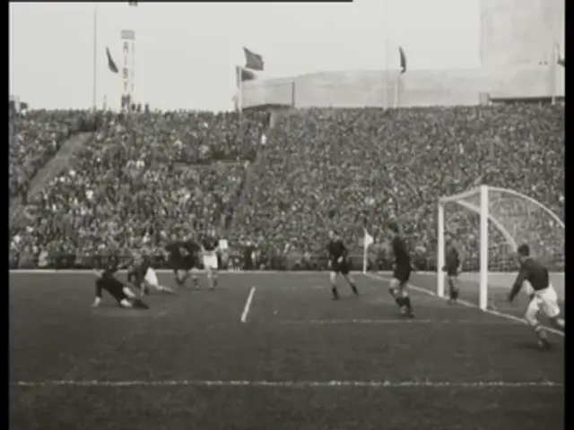 The image shows a black and white photo of a soccer game being played in a stadium, with a group of...