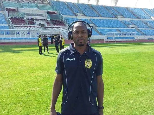 The image shows a man standing on top of a soccer field next to a stadium, wearing a headset and...