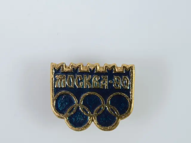 The image shows a blue and gold badge with the Olympic rings on it against a white background. The...