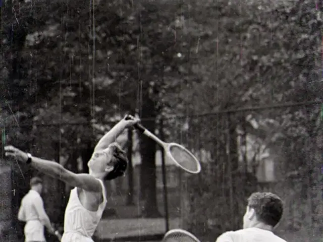 The image shows two men playing tennis on a court surrounded by trees. They are both holding...