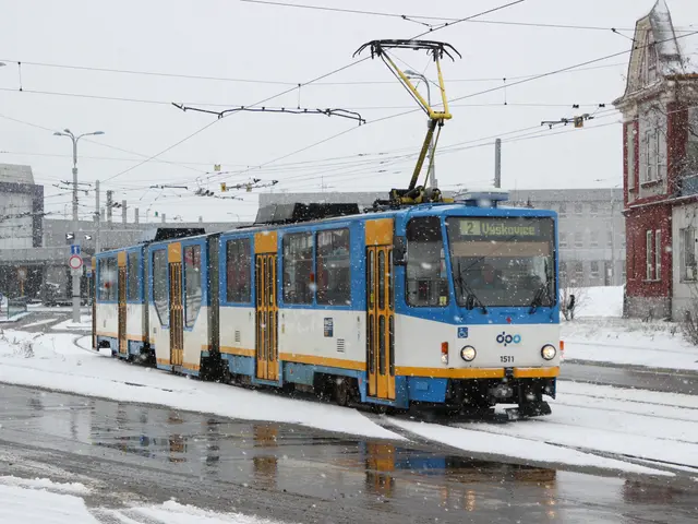The image shows a blue and yellow tram traveling down a snowy street, surrounded by buildings,...