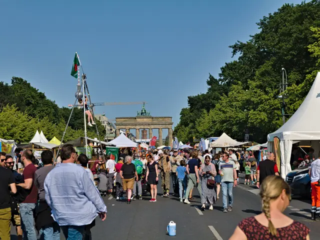The image shows a crowd of people walking down a street lined with tents, vehicles, and trees. In...
