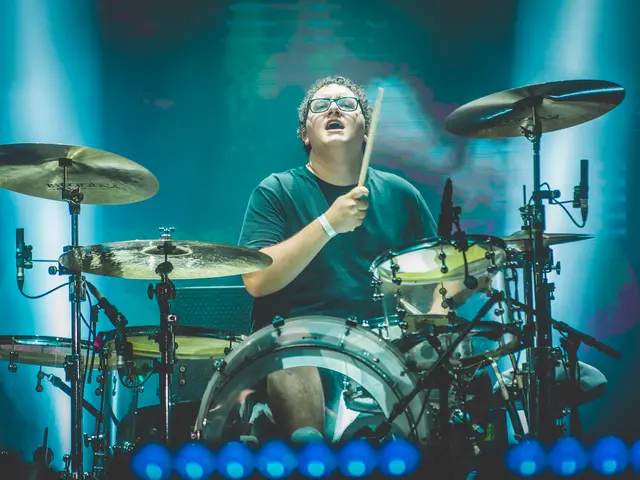 The image shows a man playing drums on stage at a concert. He is wearing a black t-shirt and...