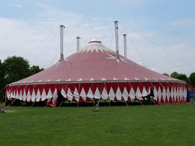 The image shows a large red and white circus tent sitting on top of a lush green field, surrounded...