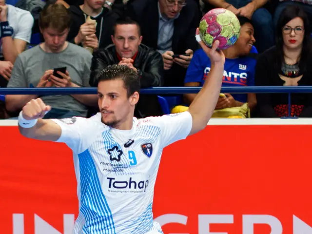 The image shows a man in mid-air, holding a handball in his hand, with a group of people sitting in...