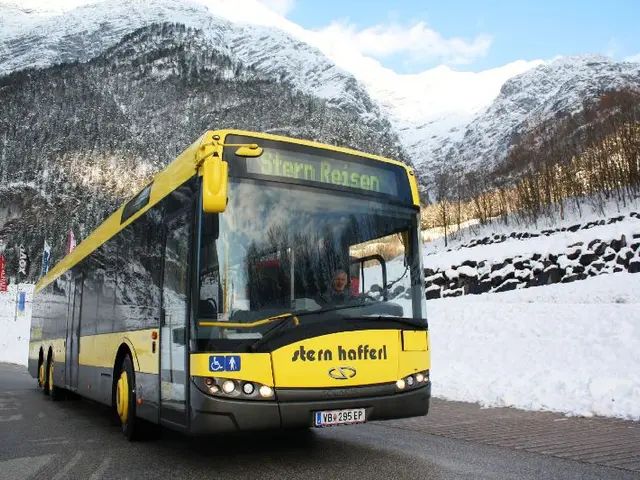 The image shows a yellow bus driving down a snowy road with mountains in the background. There are...