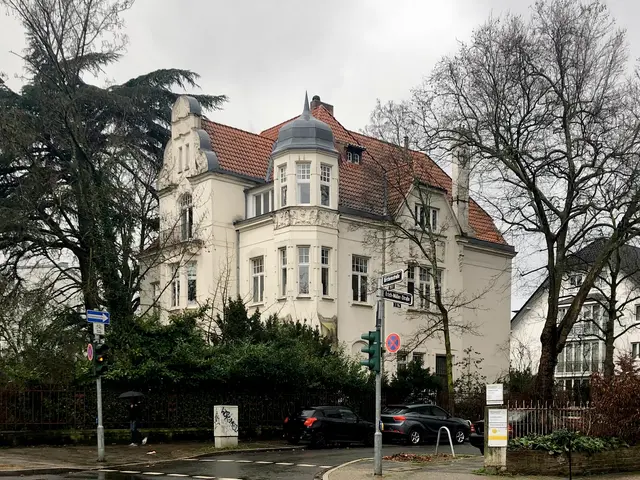 The image shows a large white building with a red roof on the corner of a street, surrounded by...